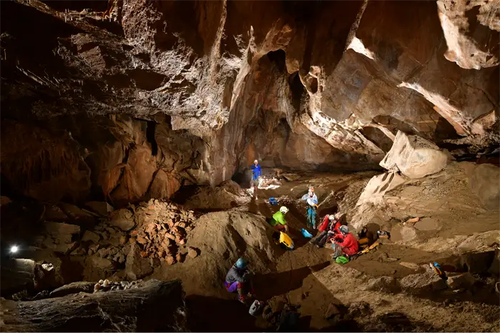 Exploratrice vêtue de rouge et portant un casque avec une lampe frontale, accroupie dans une grotte devant une formation rocheuse d'où jaillit de l'eau scintillante.
