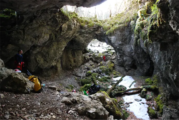 Exploratrice vêtue de rouge et portant un casque avec une lampe frontale, accroupie dans une grotte devant une formation rocheuse d'où jaillit de l'eau scintillante.