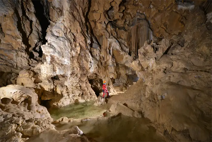 Exploratrice vêtue de rouge et portant un casque avec une lampe frontale, accroupie dans une grotte devant une formation rocheuse d'où jaillit de l'eau scintillante.