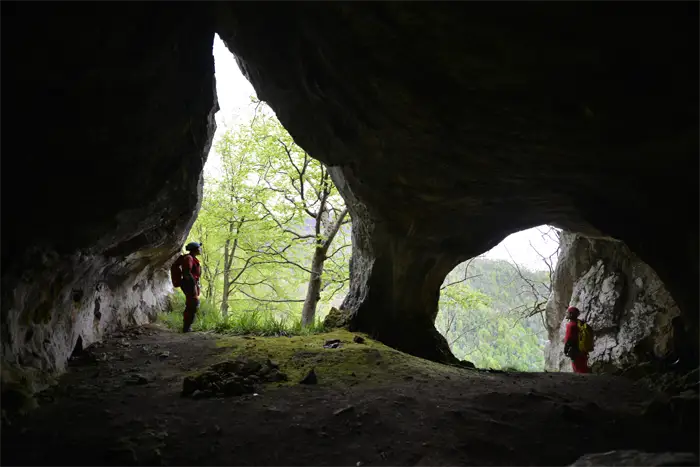 Exploratrice vêtue de rouge et portant un casque avec une lampe frontale, accroupie dans une grotte devant une formation rocheuse d'où jaillit de l'eau scintillante.