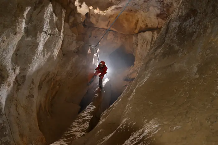 Exploratrice vêtue de rouge et portant un casque avec une lampe frontale, accroupie dans une grotte devant une formation rocheuse d'où jaillit de l'eau scintillante.