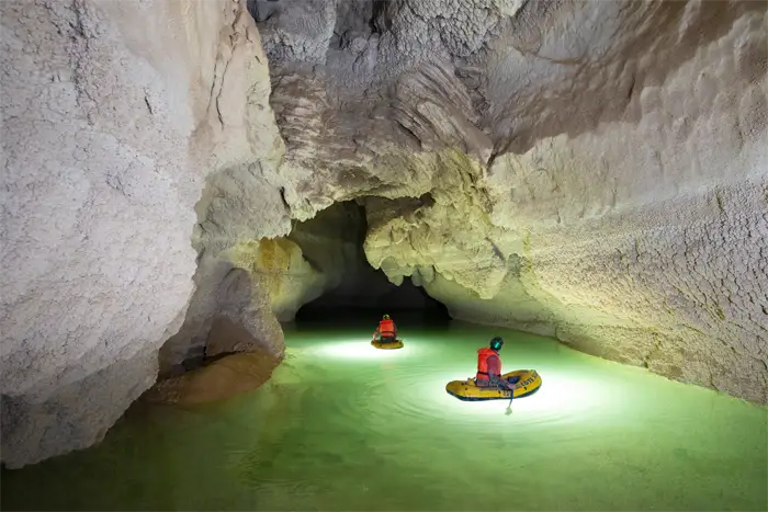 Exploratrice vêtue de rouge et portant un casque avec une lampe frontale, accroupie dans une grotte devant une formation rocheuse d'où jaillit de l'eau scintillante.