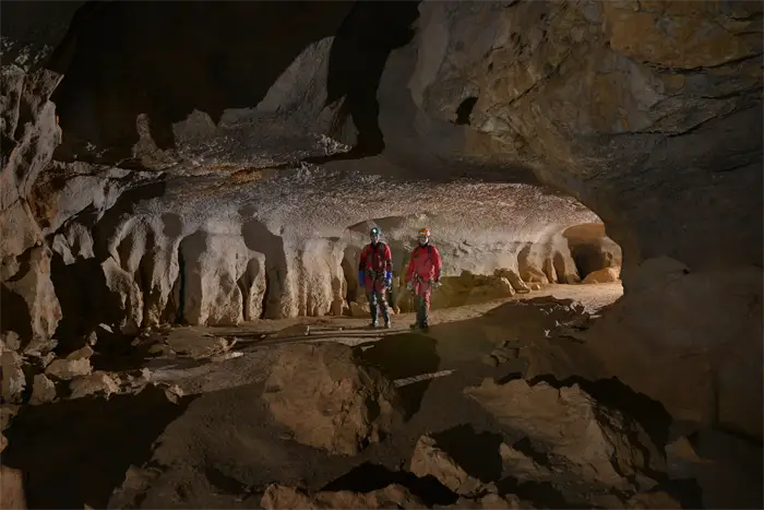 Exploratrice vêtue de rouge et portant un casque avec une lampe frontale, accroupie dans une grotte devant une formation rocheuse d'où jaillit de l'eau scintillante.