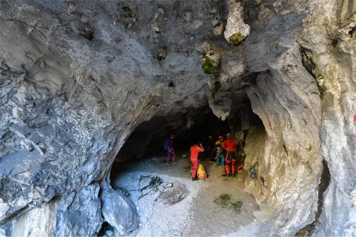 Exploratrice vêtue de rouge et portant un casque avec une lampe frontale, accroupie dans une grotte devant une formation rocheuse d'où jaillit de l'eau scintillante.