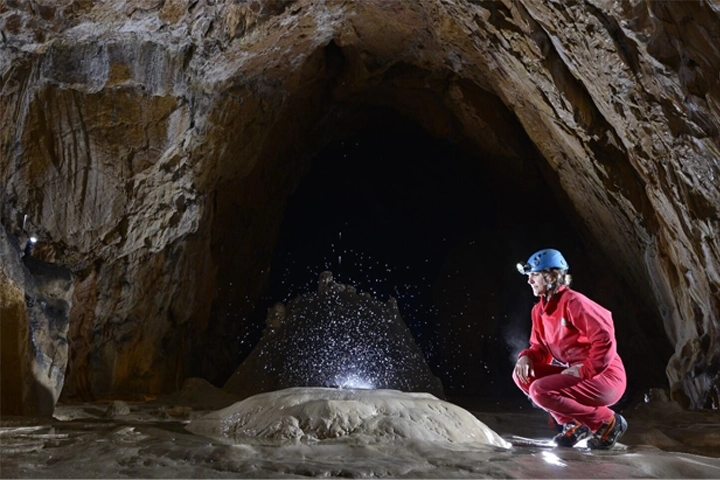 Exploratrice vêtue de rouge et portant un casque avec une lampe frontale, accroupie dans une grotte devant une formation rocheuse d'où jaillit de l'eau scintillante.